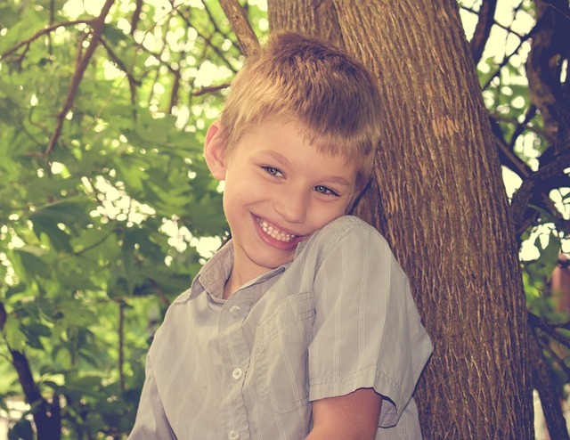 Na obrázku je smějící se chlapec s blond vlasy, modrýma očima a šedou košilí. Opírá se o strom.