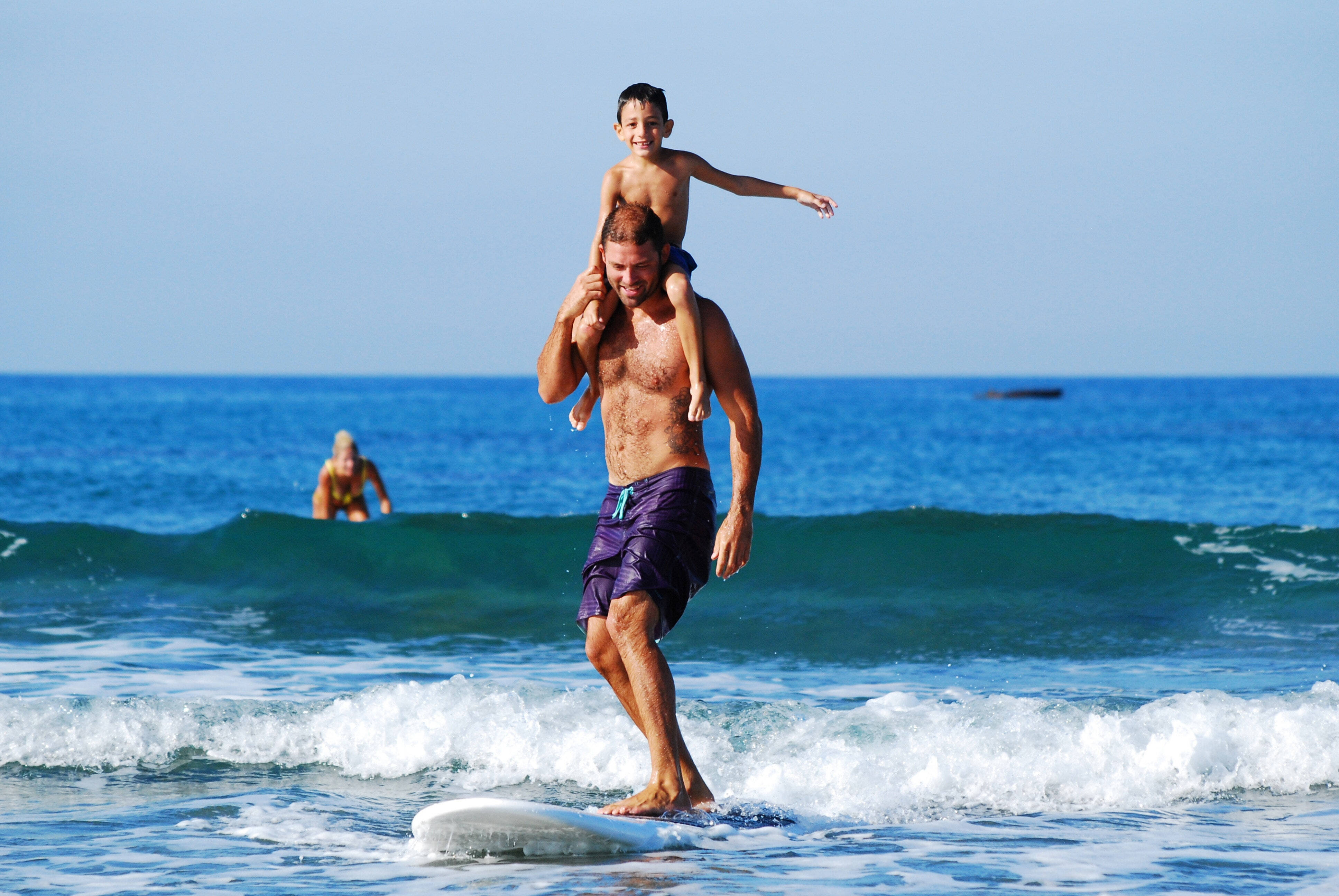 Na obrázku je otec se synem. Otec jede na surfu a syn mu sedí na ramenou. Je jasný den, moře je modré.