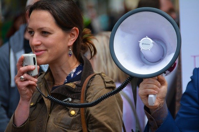Na obrázku je žena, která promlouvá na demonstraci do megafonu