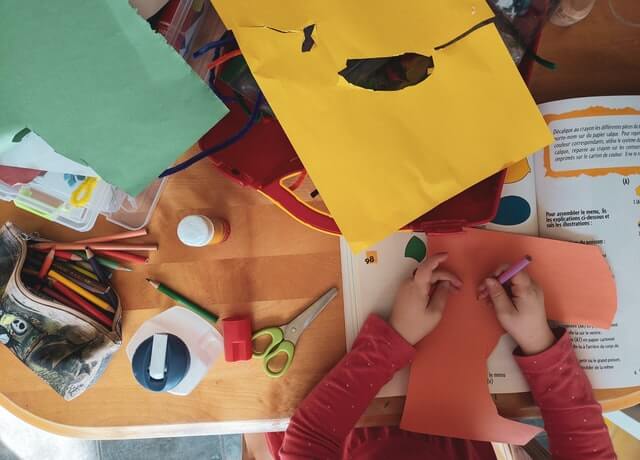 Ruce dítěte, které vystřihuje a tvoří s pomocí barevných papírů, pastelek a lepidla